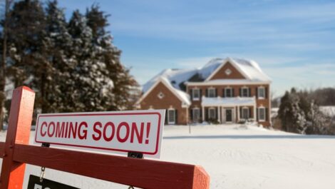 Behind a “coming soon, for sale” sign is a snow-covered home and a large, snowy yard