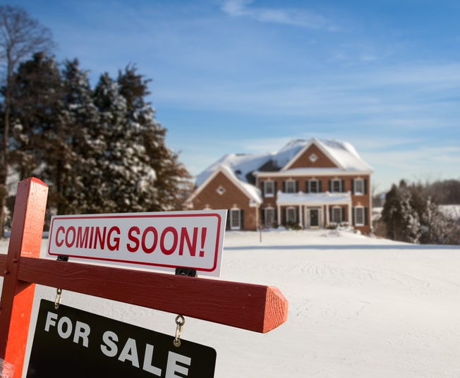 Behind a “coming soon, for sale” sign is a snow-covered home and a large, snowy yard