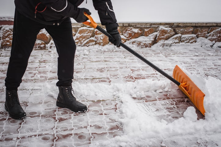 A person in a black jacket, pants, and boots is shoveling fluffy white snow from a walkway