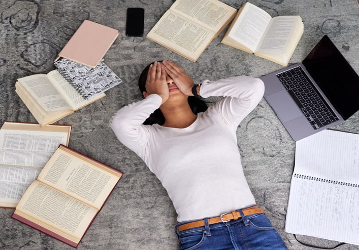 a stressed woman lies on the floor on her back with her hands over her eyes, surrounded by several open books and an open laptop