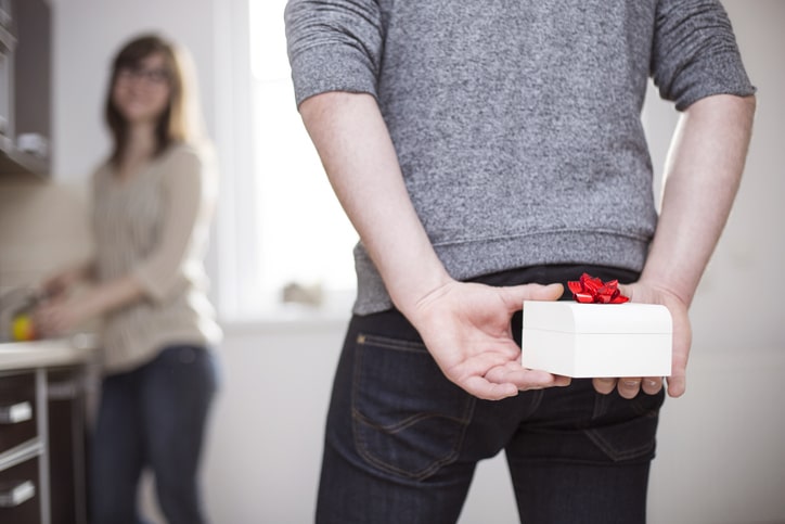 A person hides a small, white box with a red bow behind their back while a woman looks over