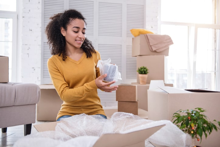 A woman is surrounded by cardboard boxes and is wrapping a mug with bubble wrap