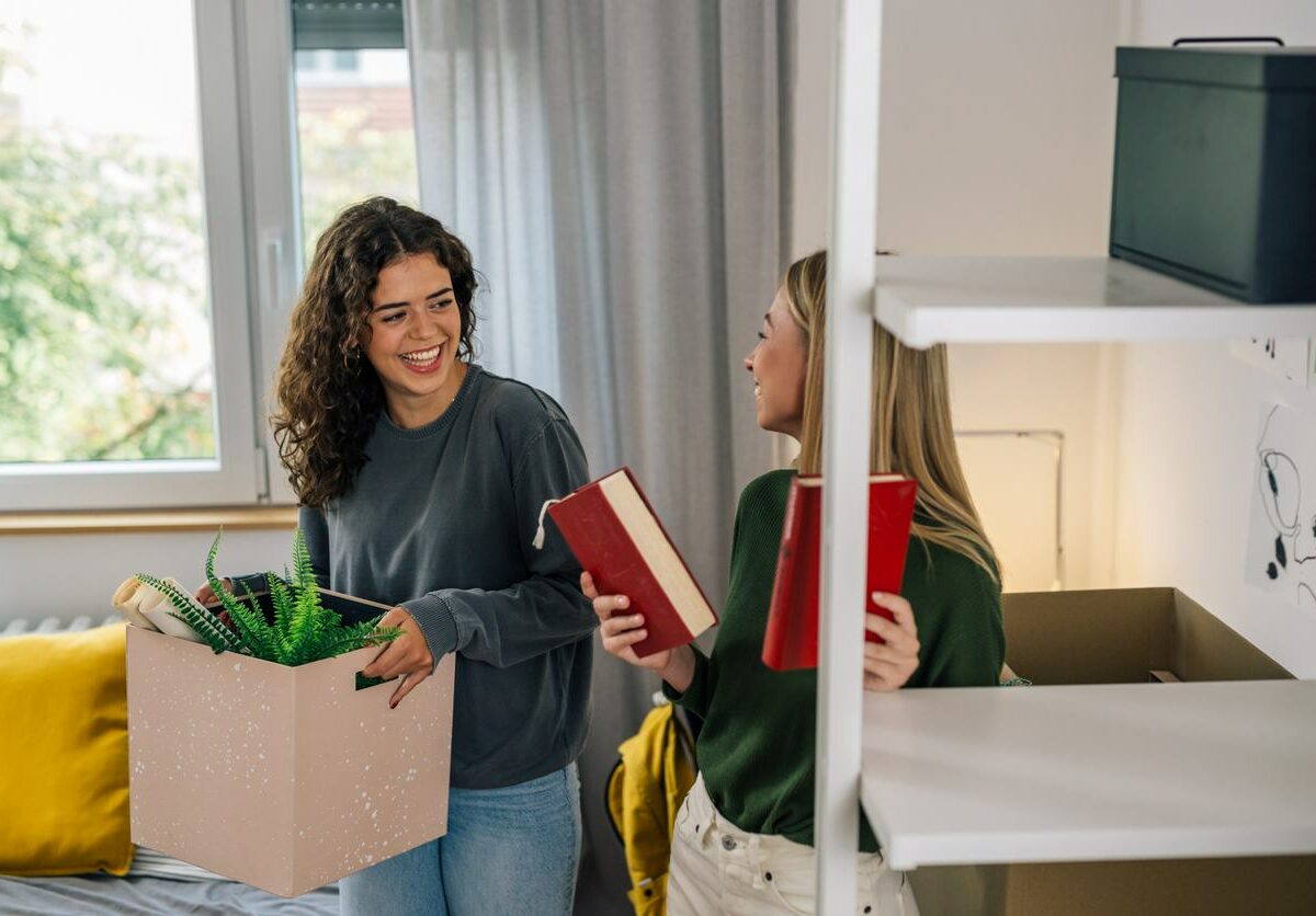 Two women carrying moving boxes out of their room