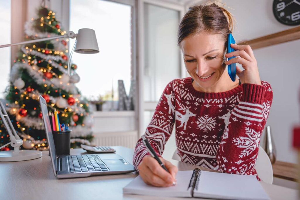 A woman holds a phone and a pen over an open book with holiday decorations nearby.