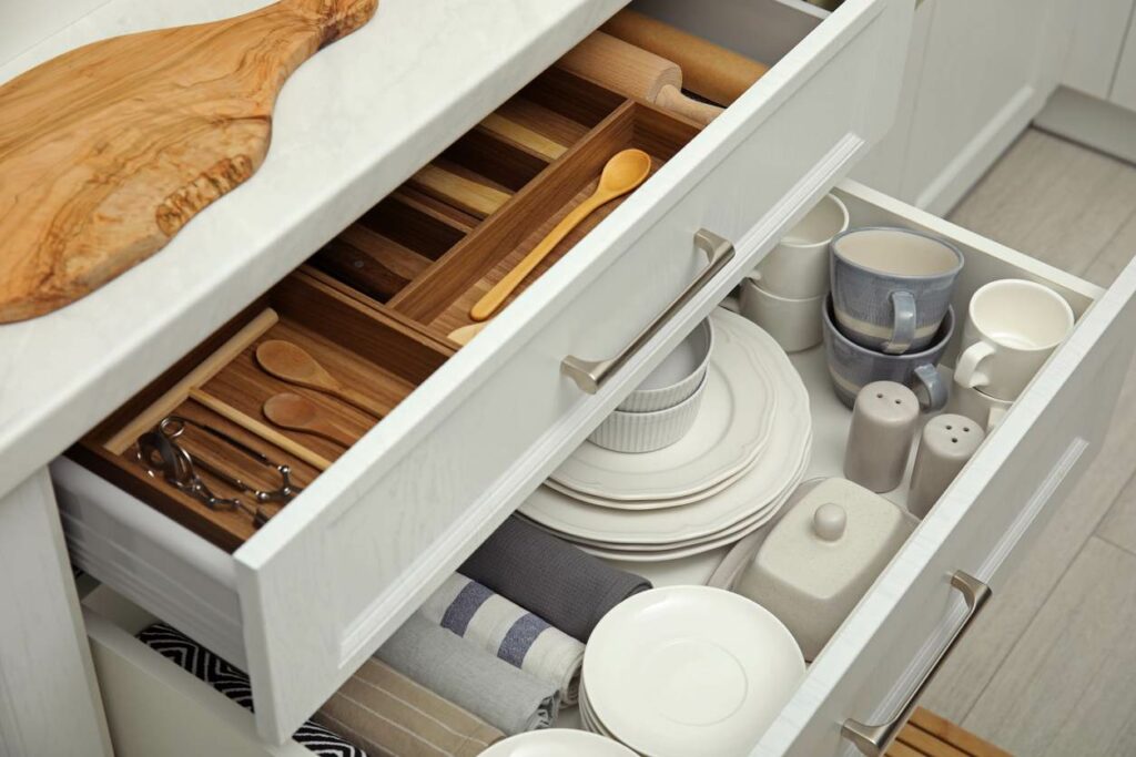 Open kitchen cabinet drawers with dishware items, utensils, and towels.