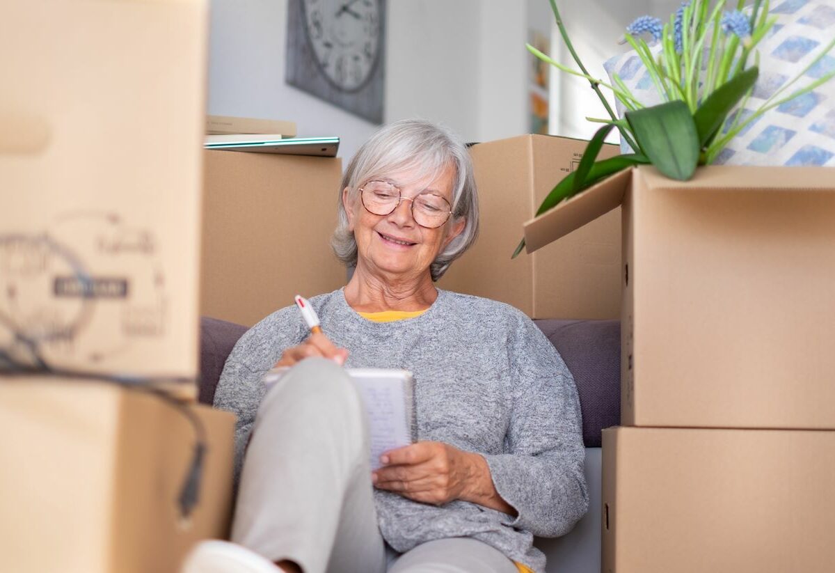 A woman writes in a notebook while sitting amid cardboard boxes during a move.