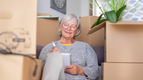 A woman writes in a notebook while sitting amid cardboard boxes during a move.