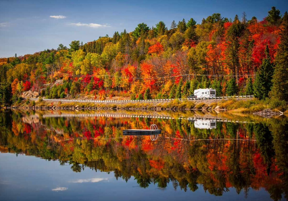 A camper driving down a road between a lake and an autumn forest