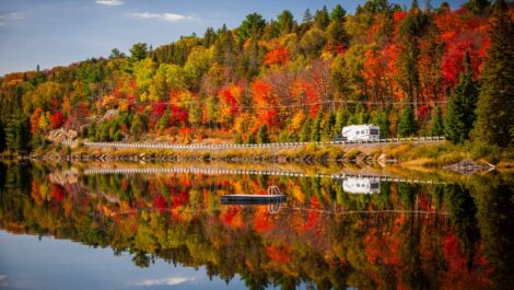 A camper driving down a road between a lake and an autumn forest