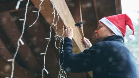 A man prepares for the holidays by hanging Christmas lights under a wooden roof.