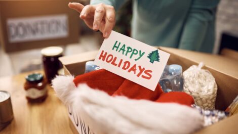 A male volunteer places a "Happy Holidays" card in a donation box filled with holiday items and canned food.