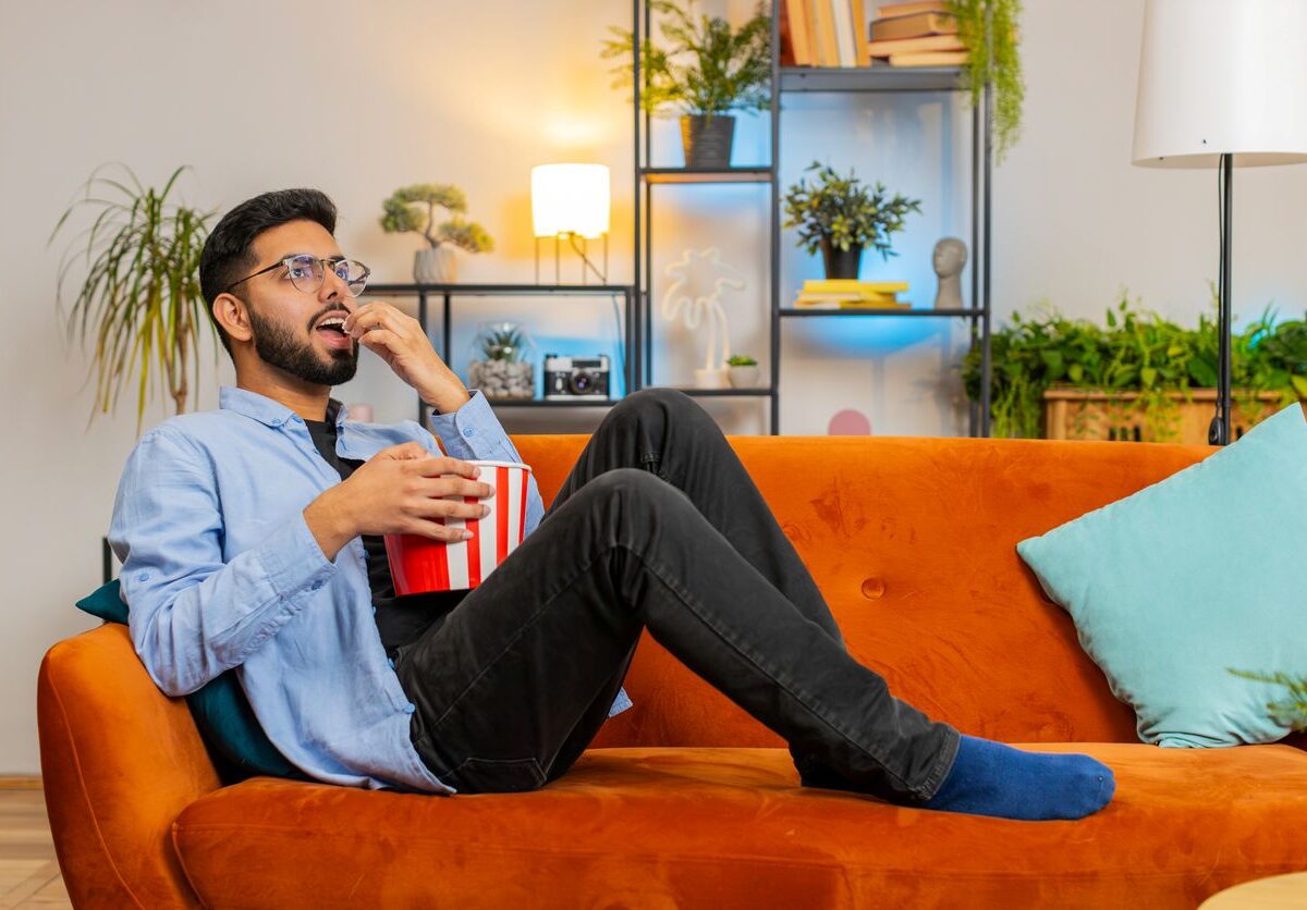 A man relaxes on an orange couch, eating popcorn and enjoying a movie, surrounded by stylish décor in his rental home.