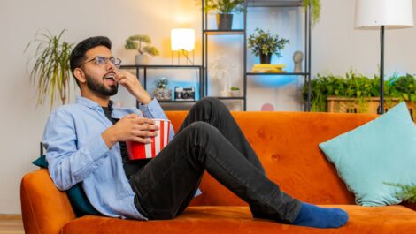 A man relaxes on an orange couch, eating popcorn and enjoying a movie, surrounded by stylish décor in his rental home.