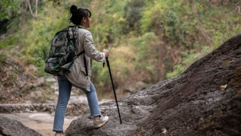 A woman hiking on a rocky trail with a trekking pole and a camouflage backpack, surrounded by a forest.