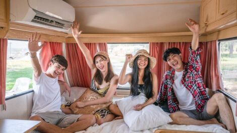 Four happy friends sitting on a bed inside an RV, smiling and raising their hands while enjoying a fun camping trip.