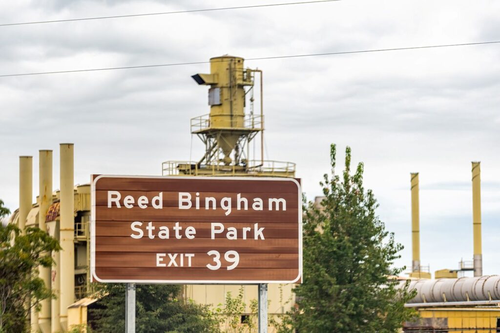 A road sign for Reed Bingham State Park at Exit 39 with industrial buildings and trees under a cloudy sky.