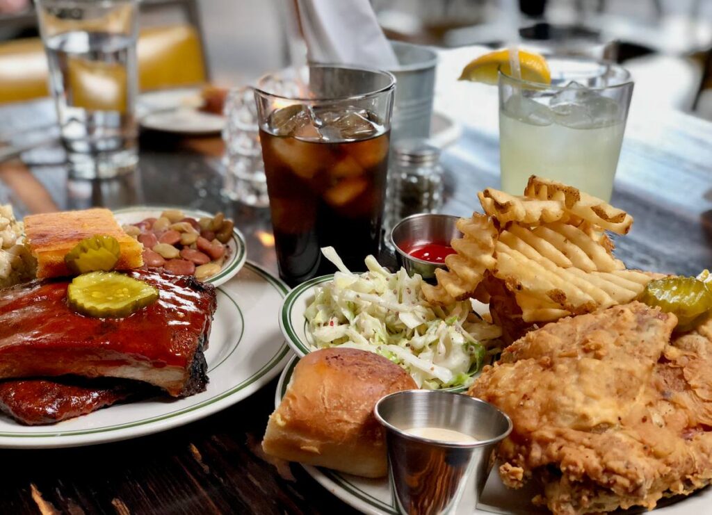 Plates of Southern food with fried chicken, ribs, coleslaw, waffle fries, and drinks served at a restaurant table.
