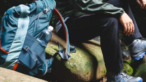 A hiker sitting on rocks in the forest with a large backpack and water bottle, wearing hiking shoes and outdoor gear.
