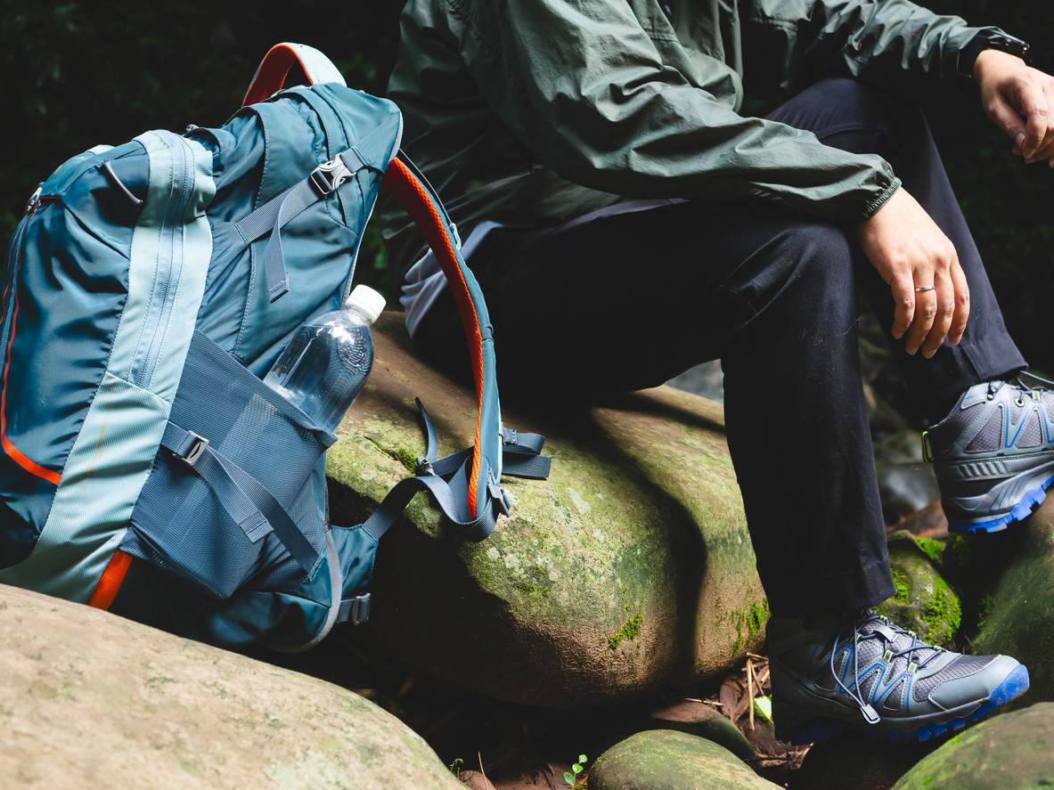 A hiker sitting on rocks in the forest with a large backpack and water bottle, wearing hiking shoes and outdoor gear.