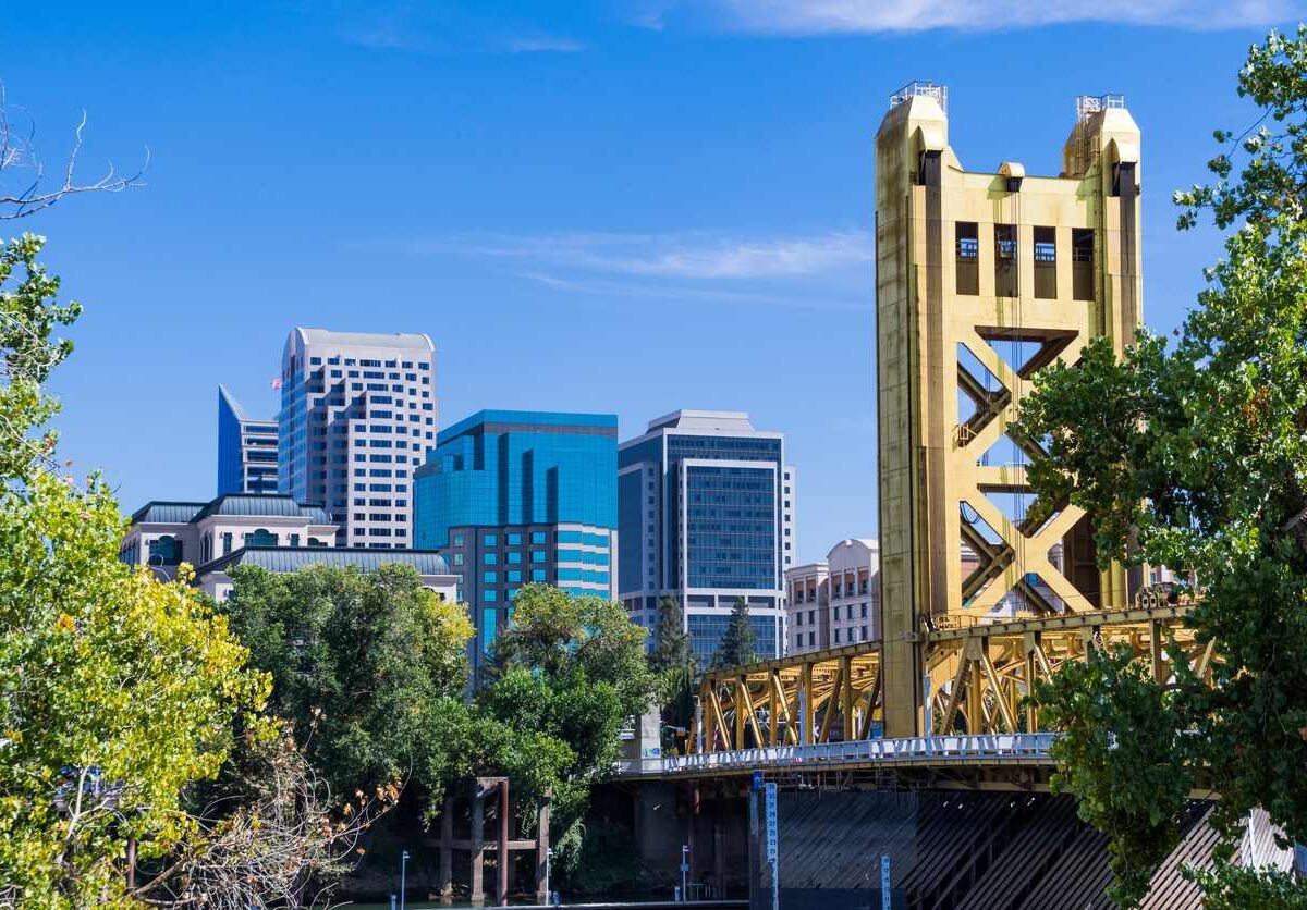 The Downtown Sacramento skyline with the iconic yellow Tower Bridge framed by green trees on a clear, sunny day.