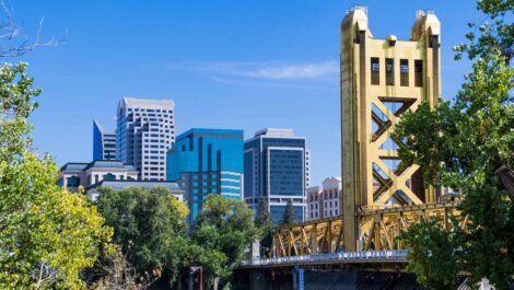 The Downtown Sacramento skyline with the iconic yellow Tower Bridge framed by green trees on a clear, sunny day.