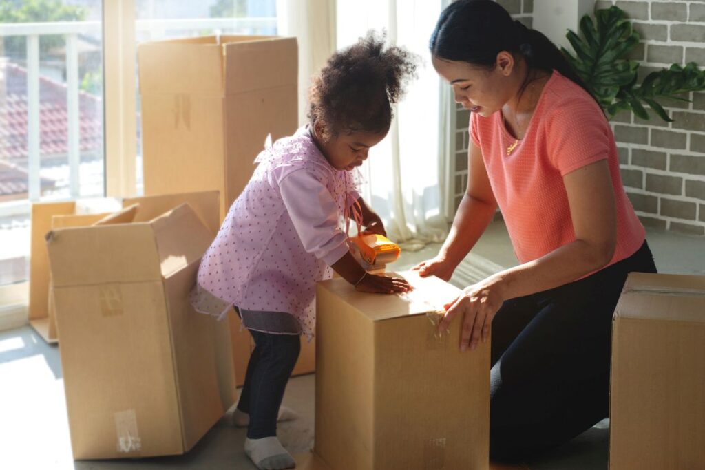 A mother and young daughter packing moving boxes together in a sunlit room, smiling and using tape to seal a box.