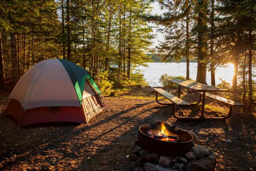A peaceful lakeside campsite with a tent, picnic table, and campfire surrounded by trees at sunset.