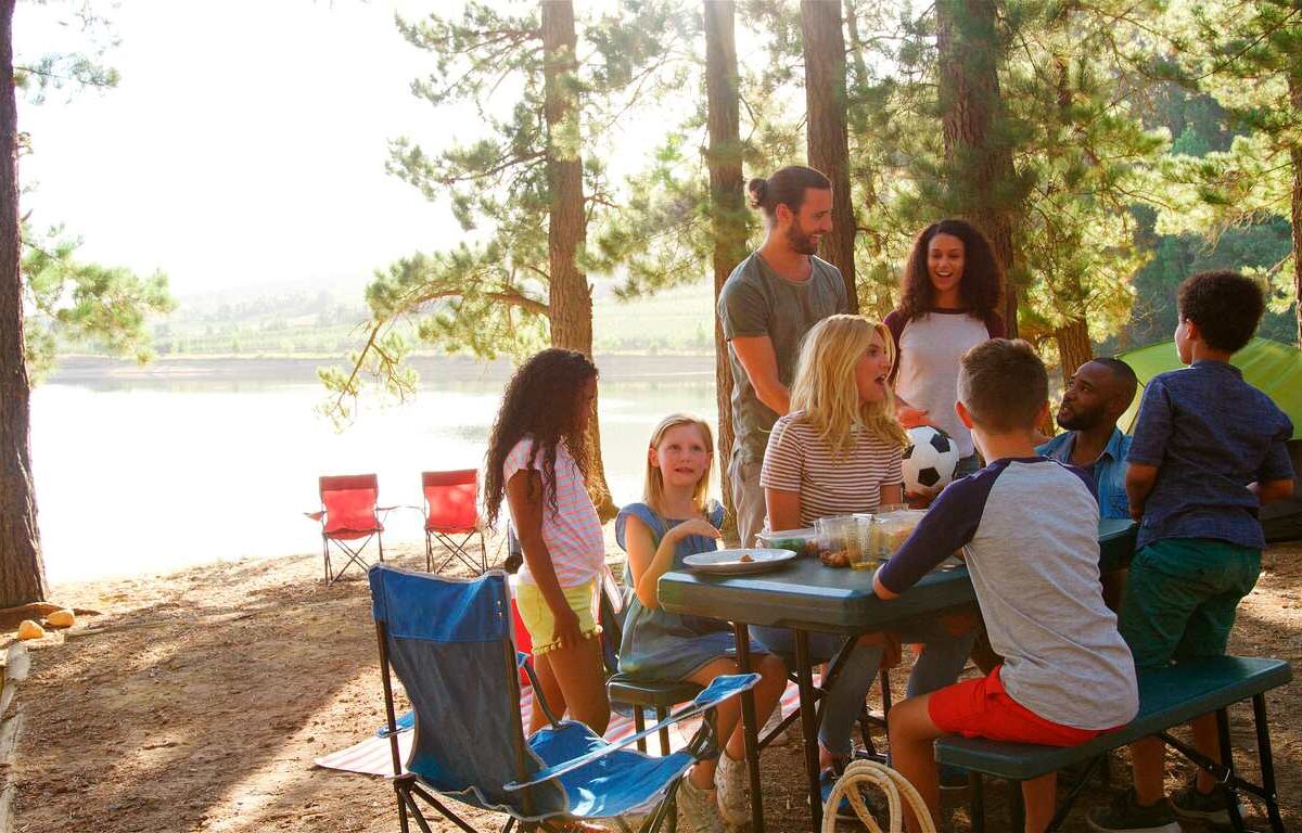 A group of friends and families enjoying a sunny camping trip together at a lakeside picnic table in the woods
