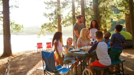 A group of friends and families enjoying a sunny camping trip together at a lakeside picnic table in the woods