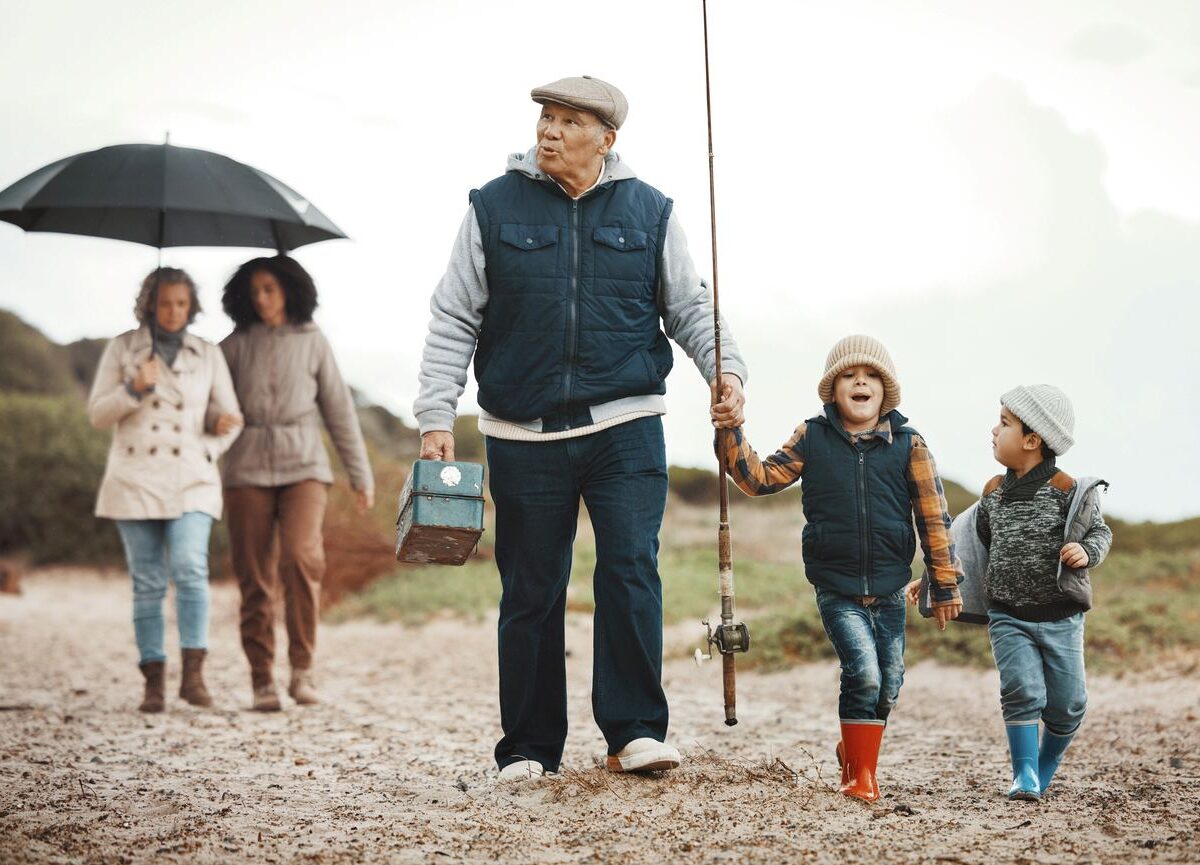 A grandfather and grandkids walk with fishing gear on a sandy path while a family follows under an umbrella.
