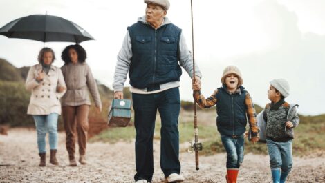 A grandfather and grandkids walk with fishing gear on a sandy path while a family follows under an umbrella.