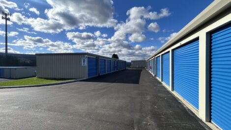 Beige metal storage building at State College with bright blue roll-up doors along the side, including one tall blue door labeled 'B' with a yellow bollard in front, situated on a gravel or asphalt lot under an overcast sky.