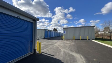 Beige metal storage building at State College with bright blue roll-up doors along the side, including one tall blue door labeled 'B' with a yellow bollard in front, situated on a gravel or asphalt lot under an overcast sky.