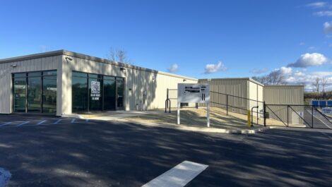 Entrance to State College Storage facility with a tall sign, wide gravel drive, chain-link perimeter fence, and storage buildings in the background.