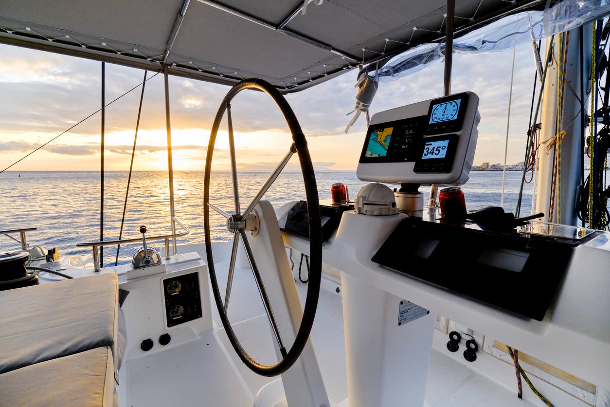 The boat’s helm with a steering wheel and navigation screens, looking out over the ocean at sunset.