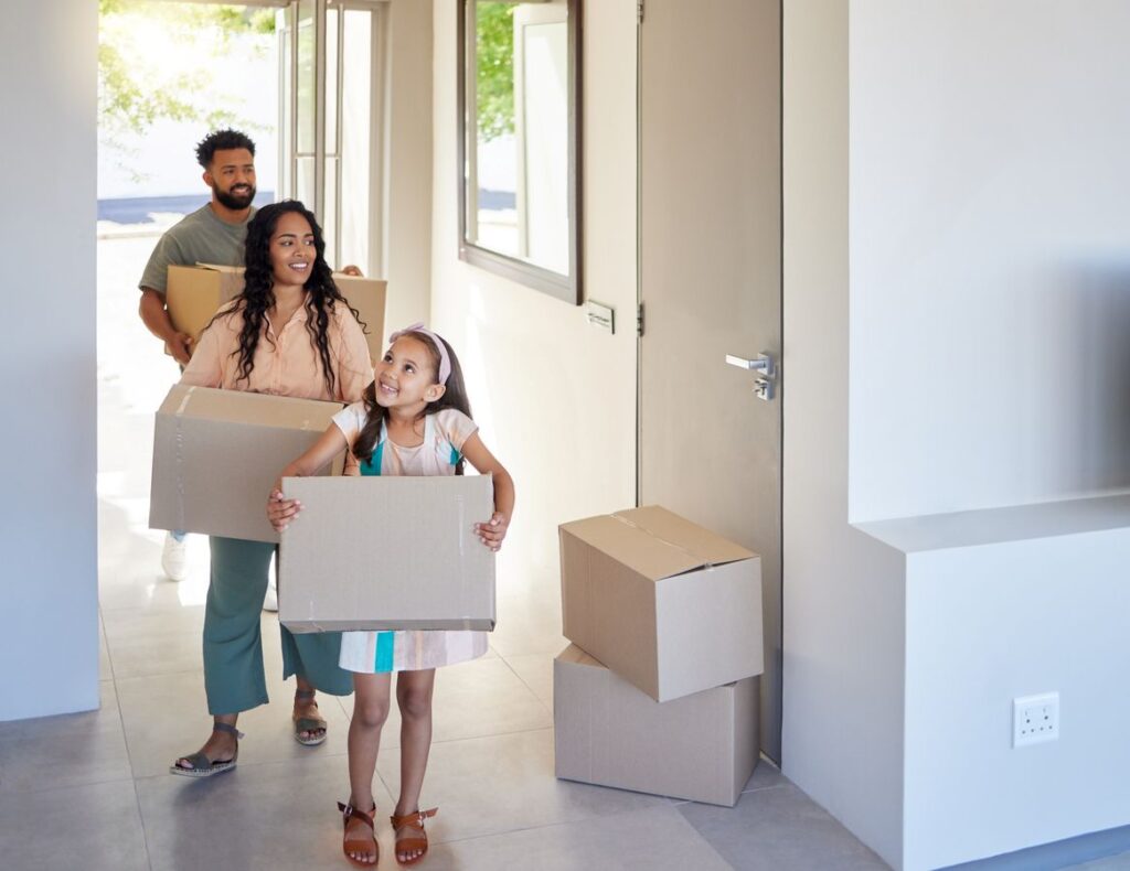 A family carrying moving boxes into their new home, smiling as they walk through the entryway together.